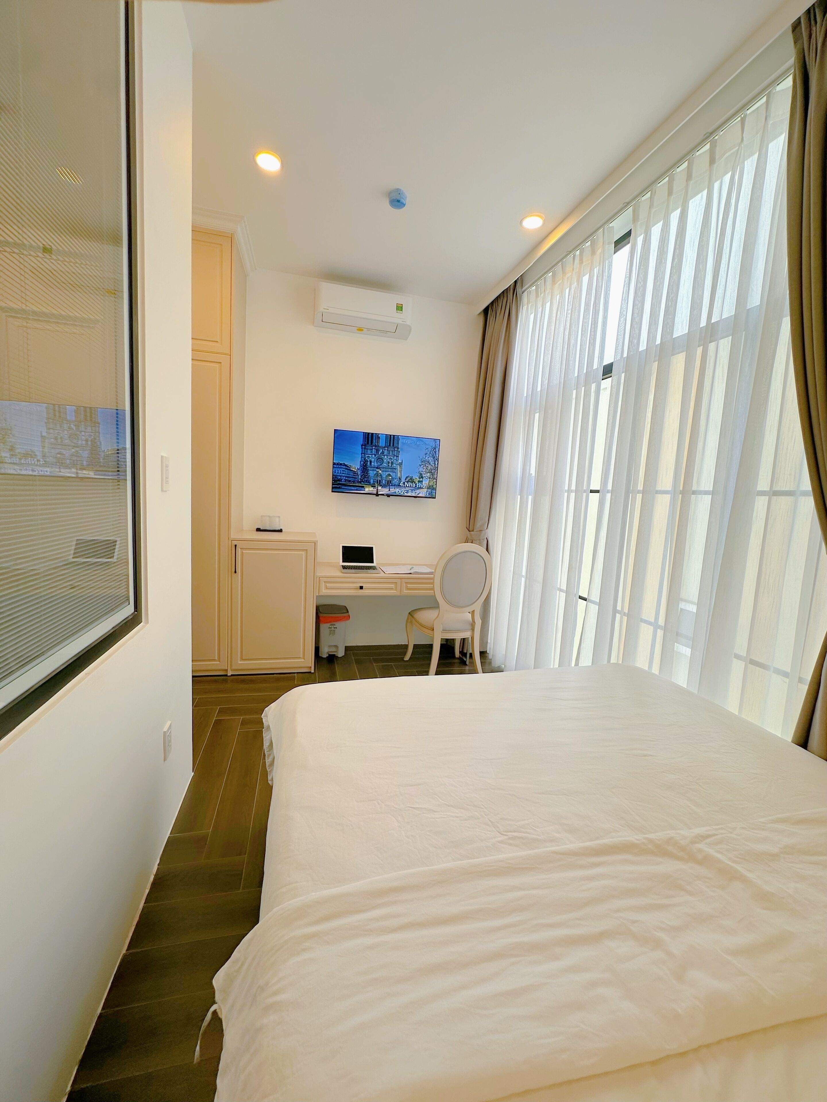 A hotel room with a bed, a desk with a laptop, a chair, a TV on the wall, and a window with curtains.