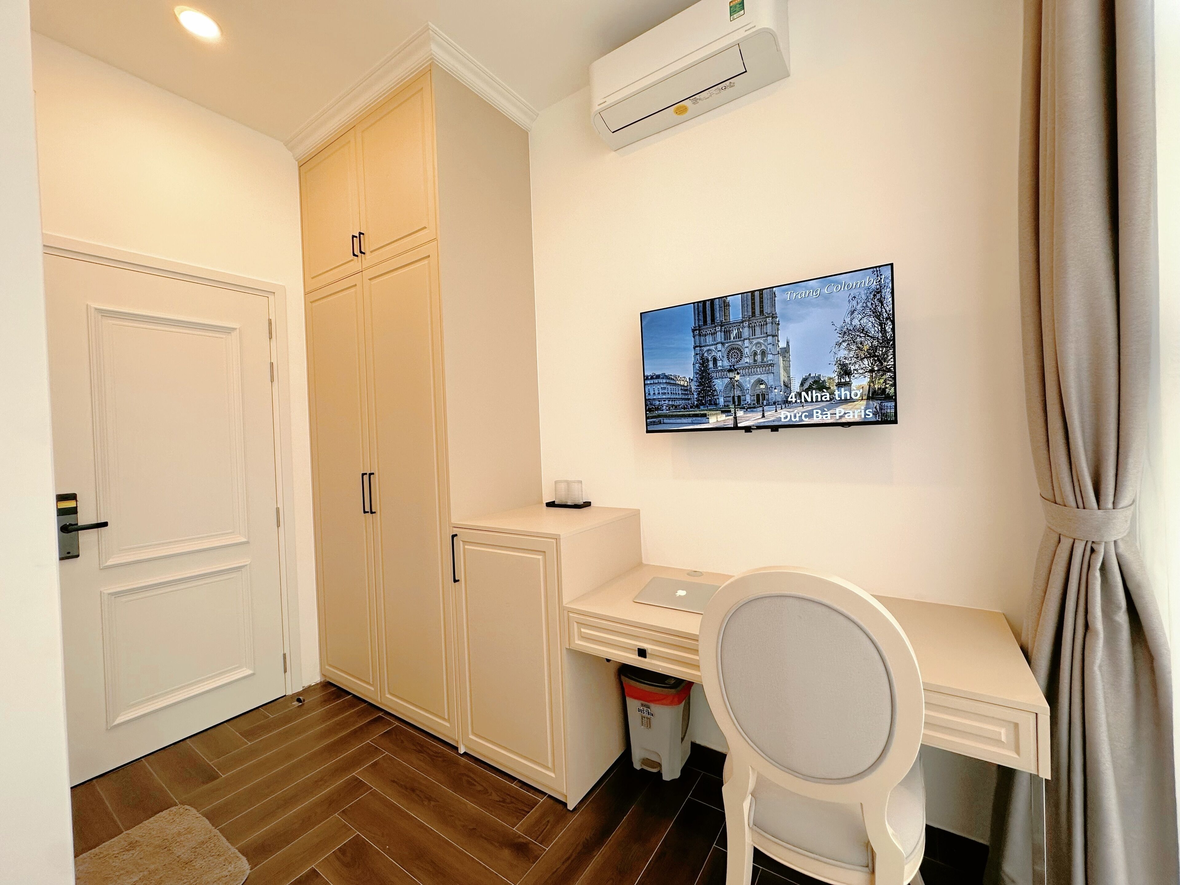 A hotel room with a white desk, a chair, a television mounted on the wall, and a wardrobe.