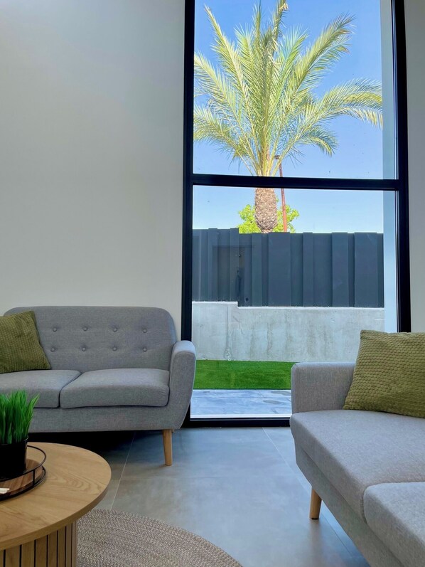 A modern living room with two grey sofas, a wooden coffee table, and a large window offering a view of a palm tree and a fenced yard.