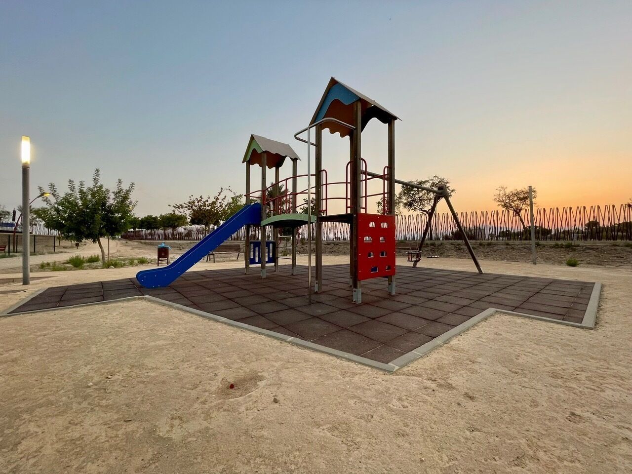 Ein Spielplatz mit einer blauen Rutsche und Klettergerüsten, umgeben von einem Zaun und einer Straßenlaterne.