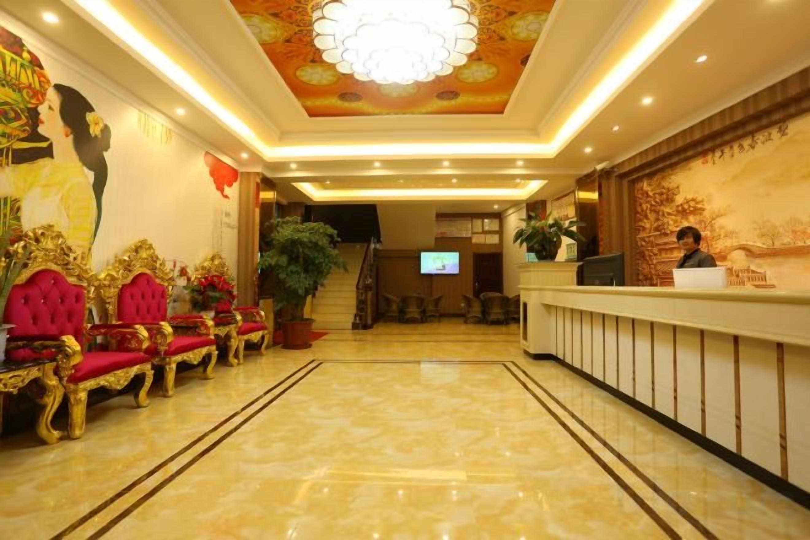 A hotel lobby with a reception desk, red upholstered chairs, a large chandelier, and a television screen.