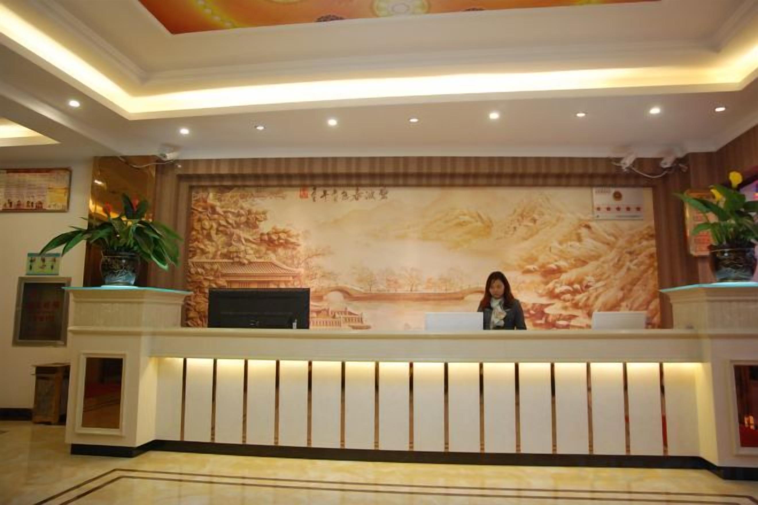 A hotel reception desk with a receptionist, a large mural, and potted plants.