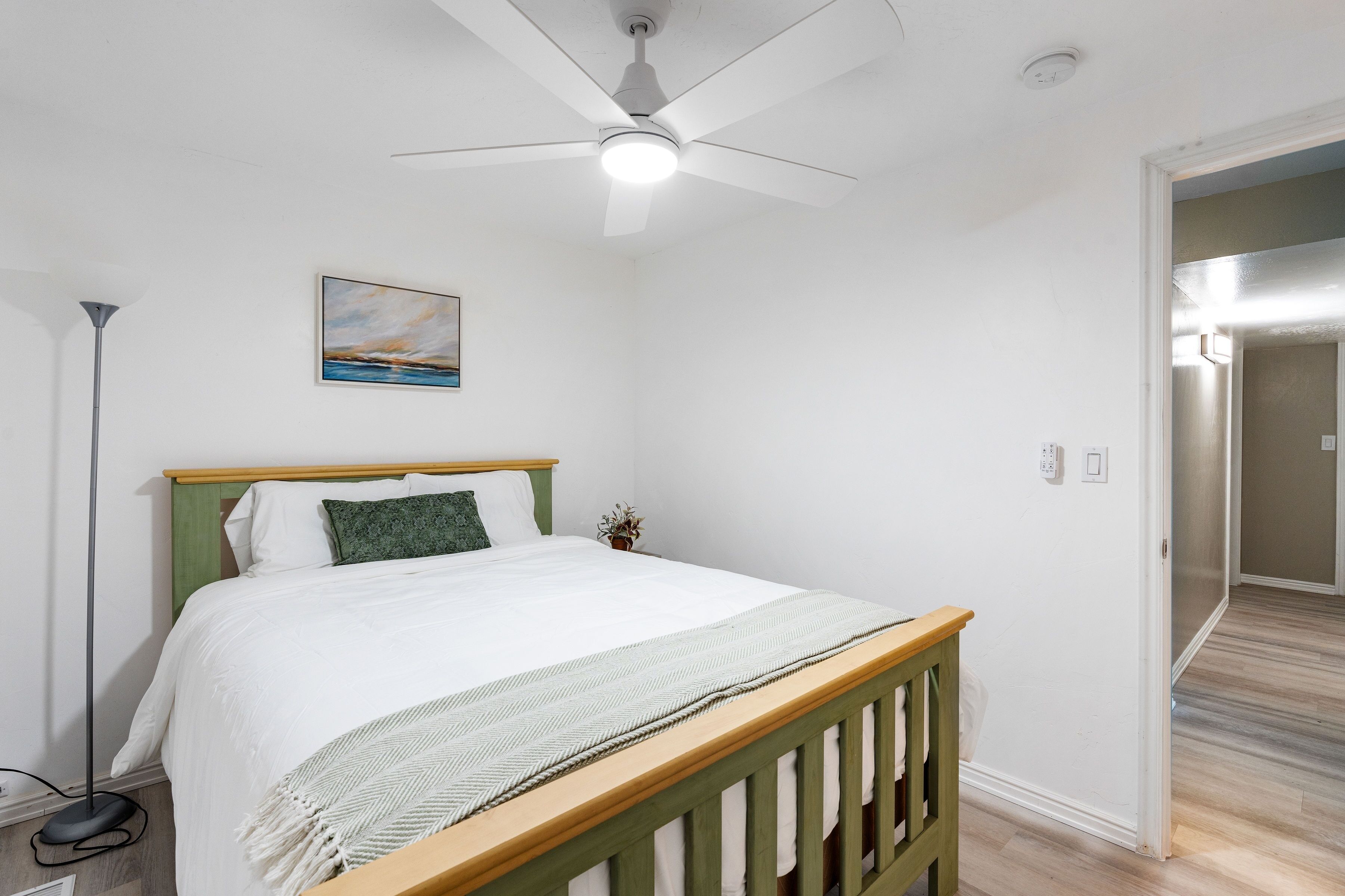 A bedroom with a bed, a ceiling fan, a painting, and a floor lamp.