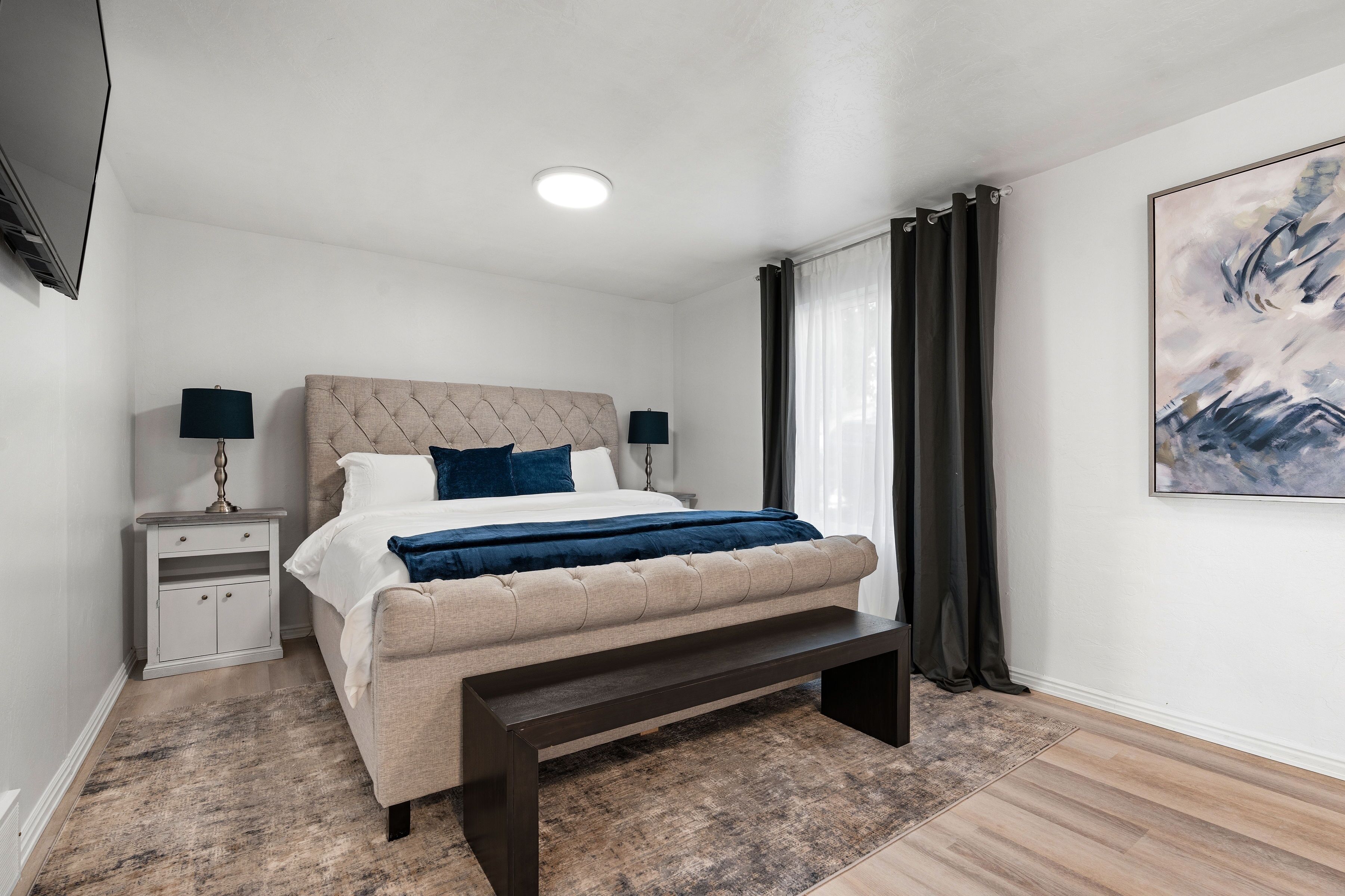 A modern bedroom with a tufted headboard, a bed with white and blue bedding, a bedside table with lamps, a bench at the foot of the bed, and a large abstract painting on the wall.