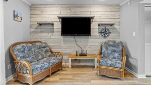 Uma sala de estar com dois sofás de vime, uma TV de tela plana fixada em uma parede de madeira e uma pequena mesa de madeira.
