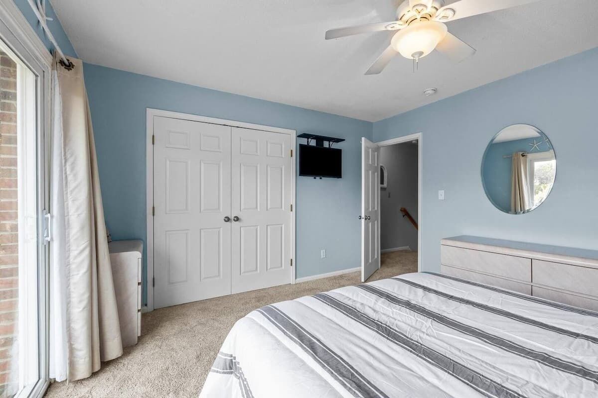 A bedroom with a bed, a ceiling fan, a door, and a wall-mounted TV.