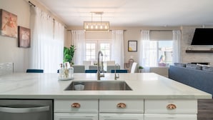 Une cuisine moderne dotée d’un îlot central, d’un évier en acier inoxydable et de meubles blancs. On y trouve également un coin repas avec des chaises bleues et un canapé gris.