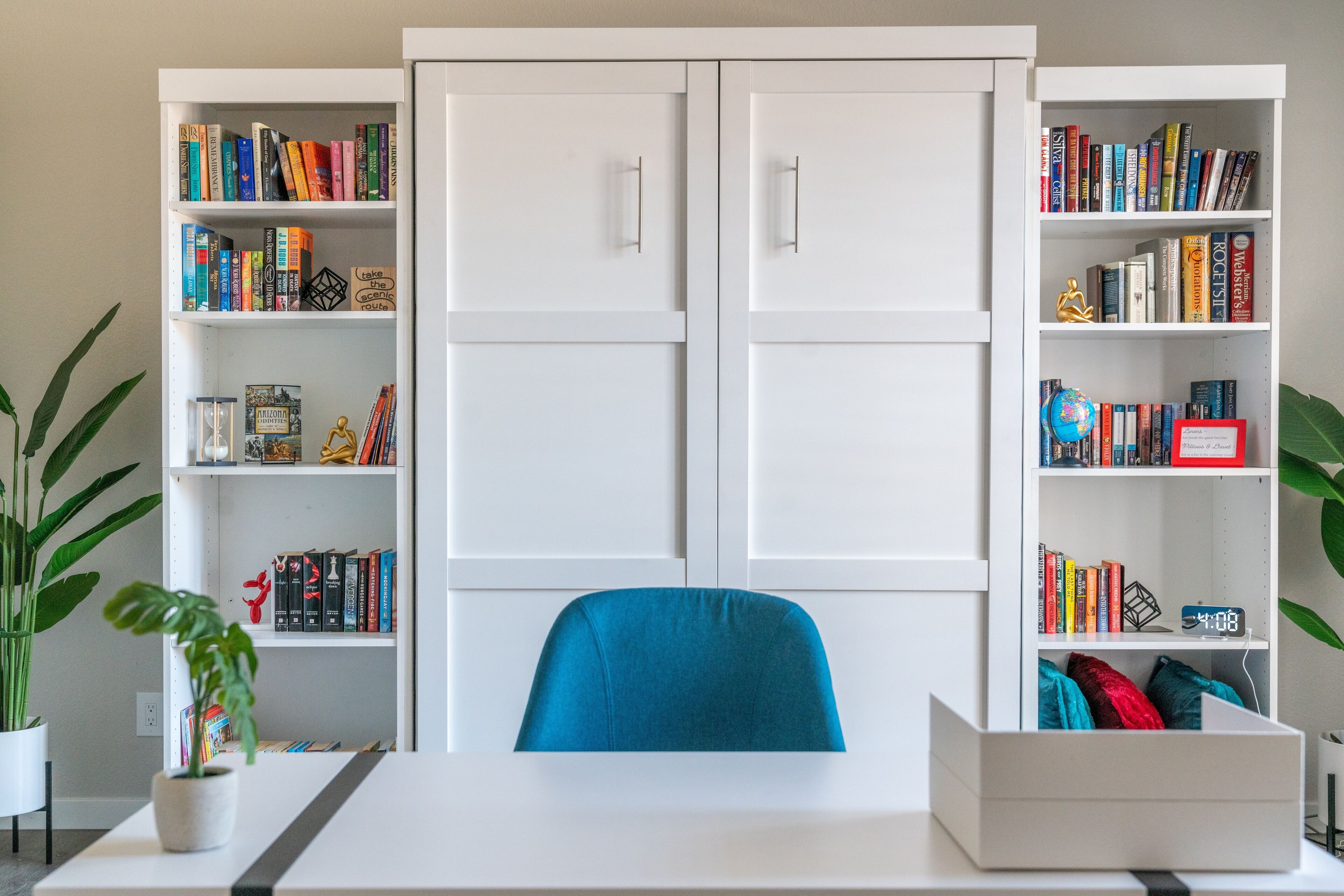 A room with a white desk, a blue chair, and two white bookshelves filled with books and decorative items.