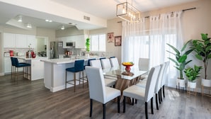 Une cuisine moderne avec des meubles blancs, un îlot central et un coin repas comprenant une table en verre et des chaises blanches.