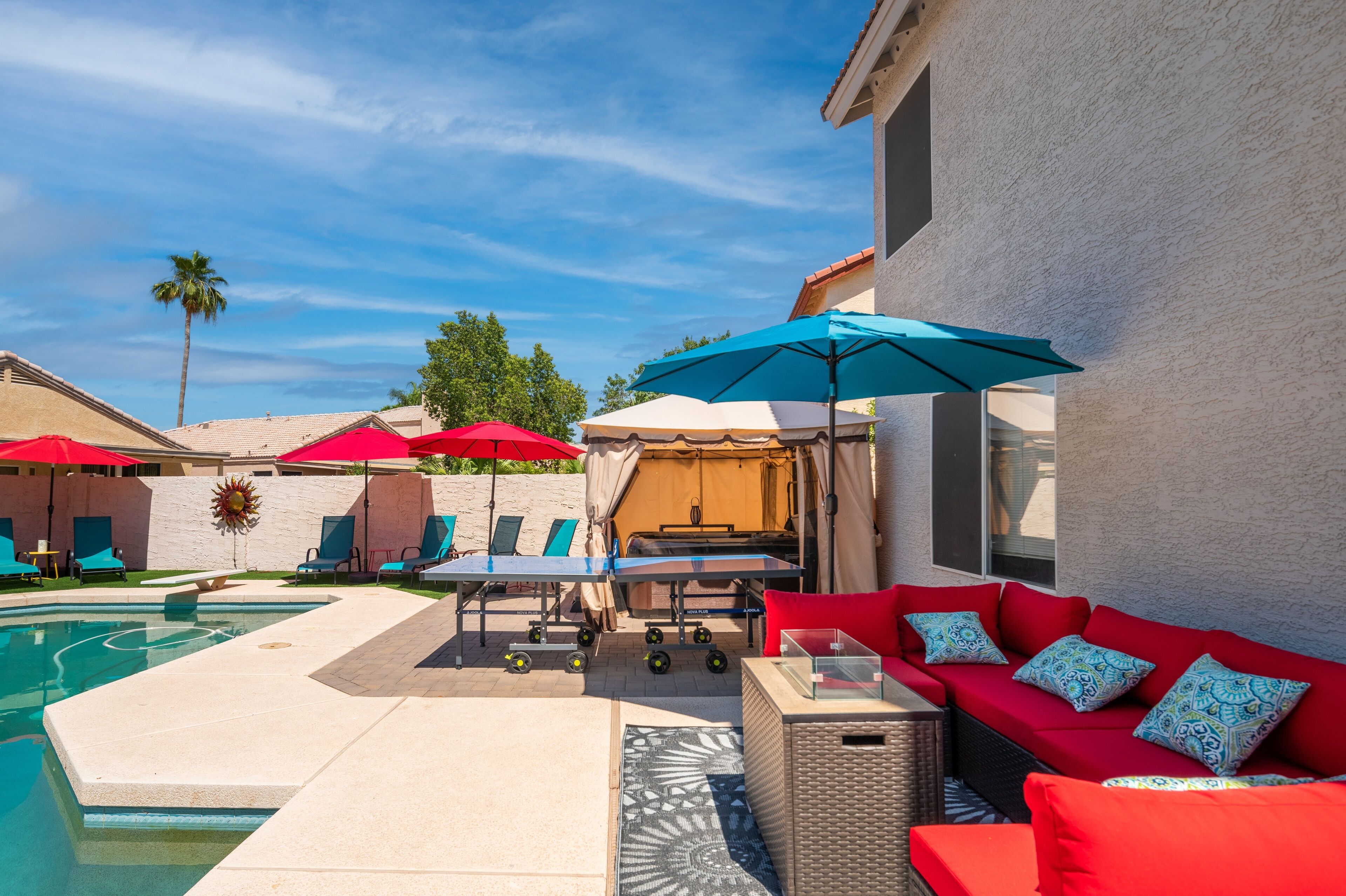 A poolside area with lounge chairs, umbrellas, and a red sofa with patterned cushions.