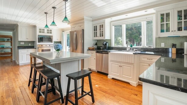 A spacious kitchen with white cabinetry, a central island with stools, stainless steel appliances, and pendant lighting. - 5BR Cape Cod Retreat, Short Walk to Beach (West Yarmouth)