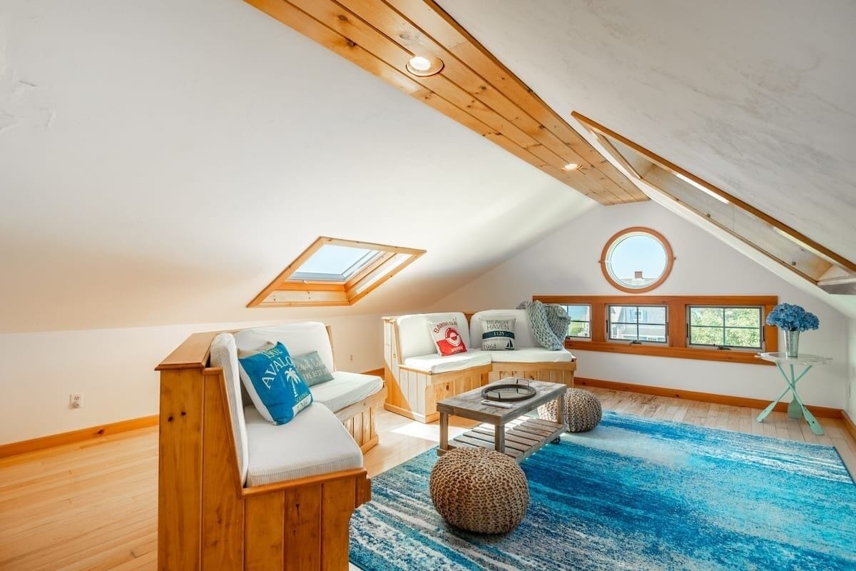 Une chambre mansardée chaleureuse, avec un plafond en pente, des poutres apparentes, un puits de lumière, une fenêtre ronde et un tapis à rayures bleues.