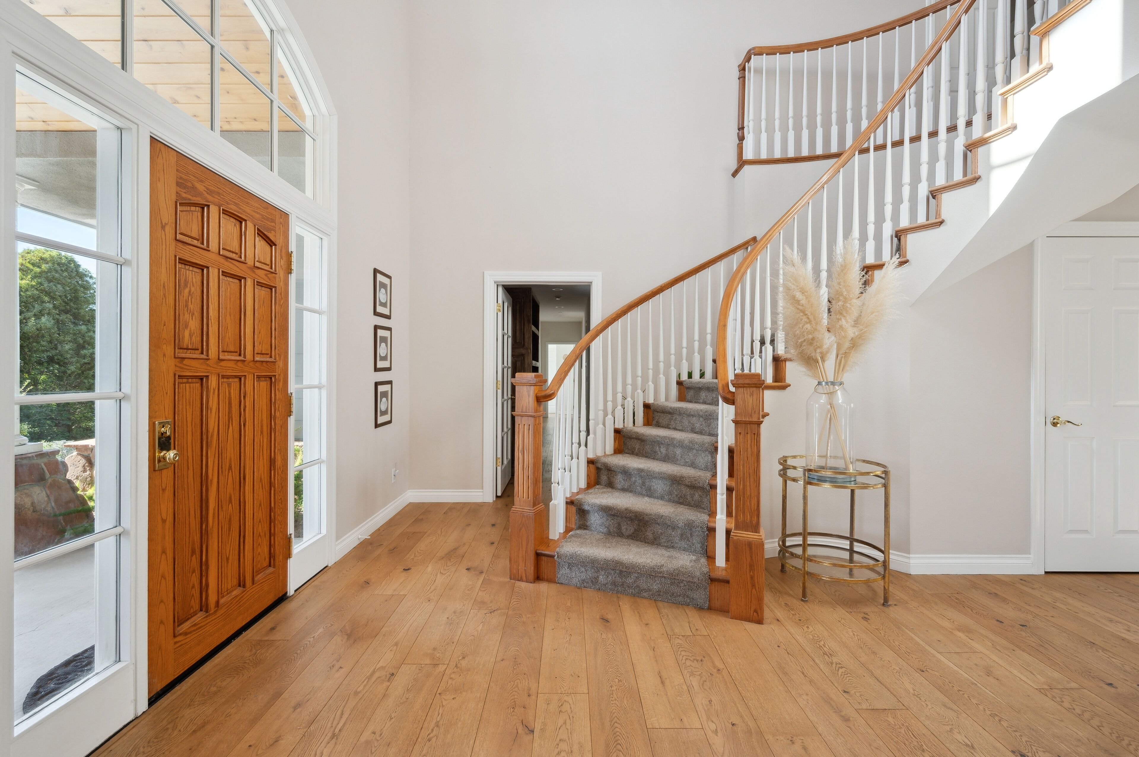A spacious hallway with a wooden staircase, large windows, and a door leading outside.