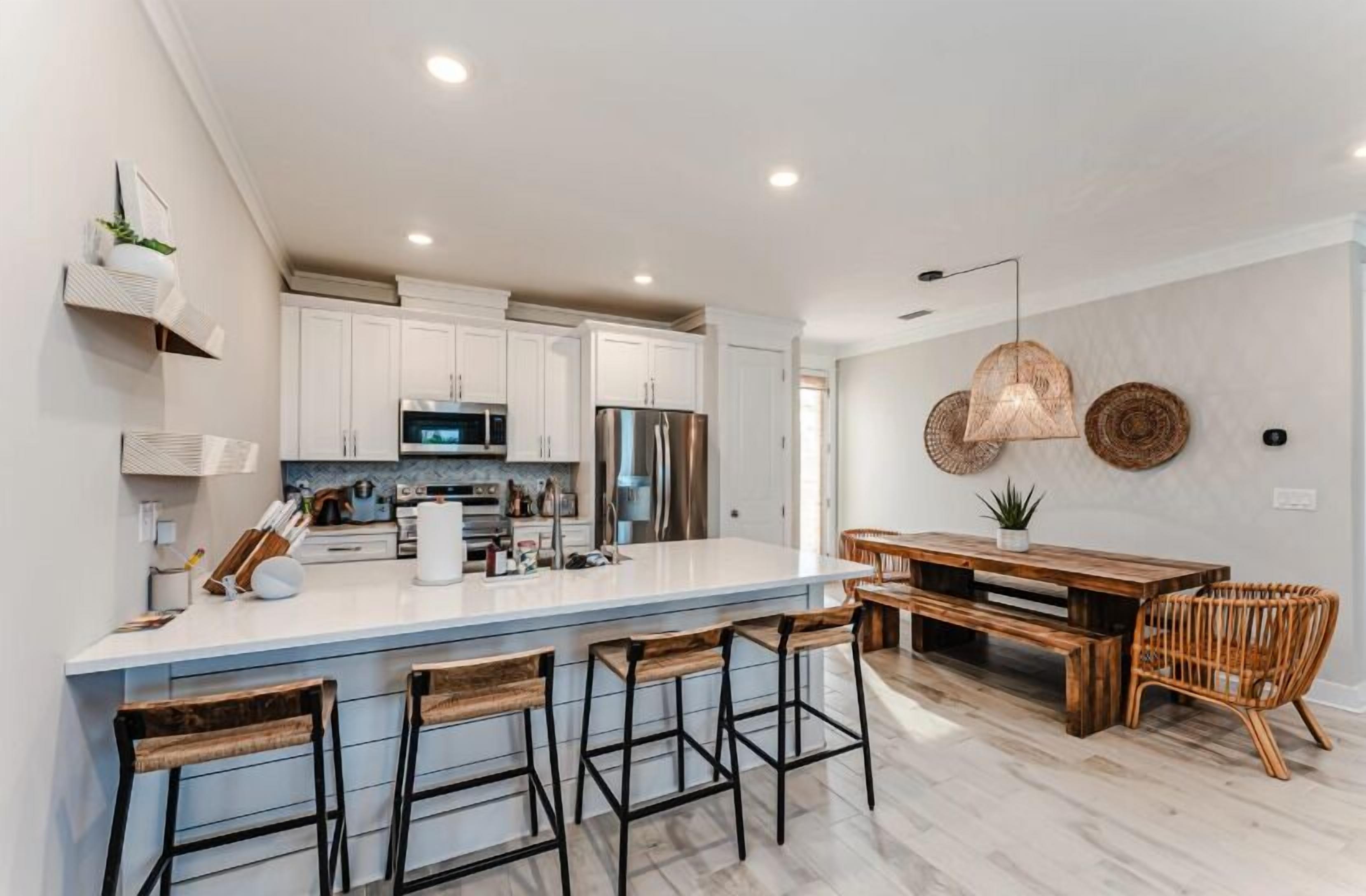 Una cocina moderna con armarios blancos, electrodomésticos de acero inoxidable, una isla grande con taburetes de bar y un comedor con mesa y sillas de madera.