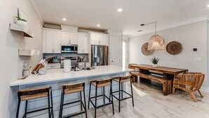A modern kitchen with white cabinets, stainless steel appliances, a large island with bar stools, and a dining area with a wooden table and chairs. - Port St Joe Beach House Pool 5BR Sleeps 12 by RedAwning (Port St. Joe)