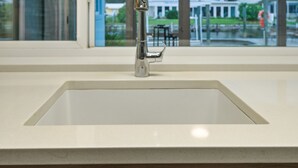 Una cocina moderna con un grifo de acero inoxidable, encimera blanca y vistas a una casa a través de una puerta de vidrio.