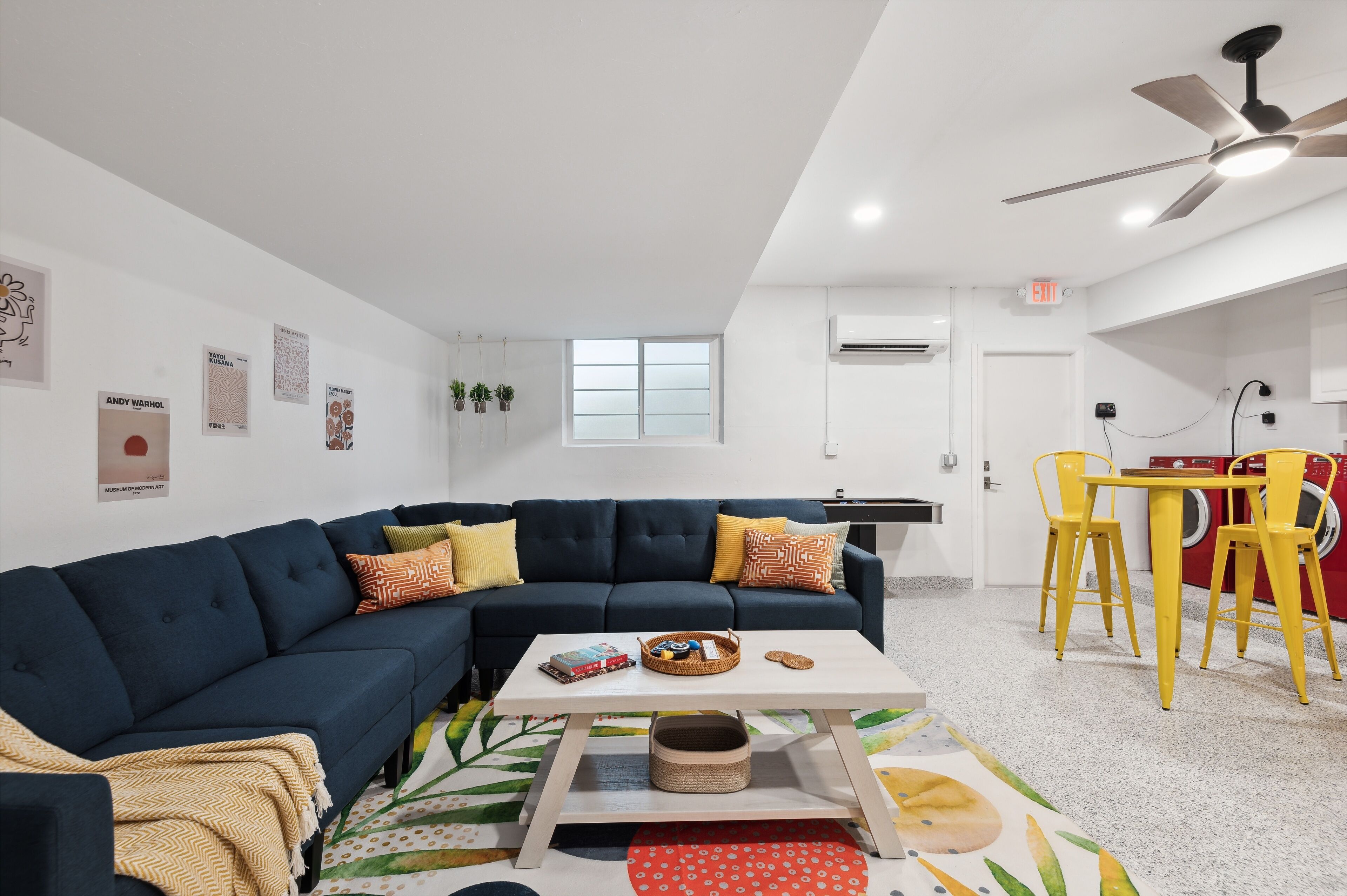 Un salon moderne avec un canapé bleu foncé, une table basse blanche et des chaises jaunes. On y trouve une fenêtre à vitres dépolies, un climatiseur et un ventilateur de plafond.