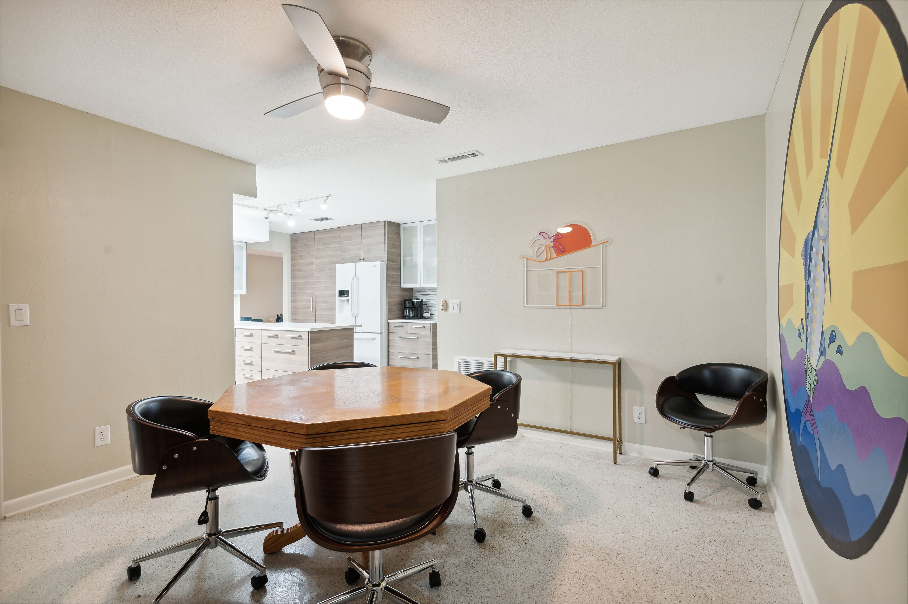 A conference room with a wooden table, four black office chairs, a ceiling fan, and a kitchen in the background.