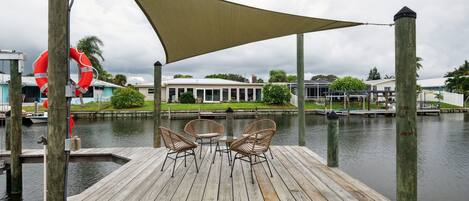 A wooden deck with two wicker chairs and a table, overlooking a calm body of water with a lifebuoy attached to a pole.
