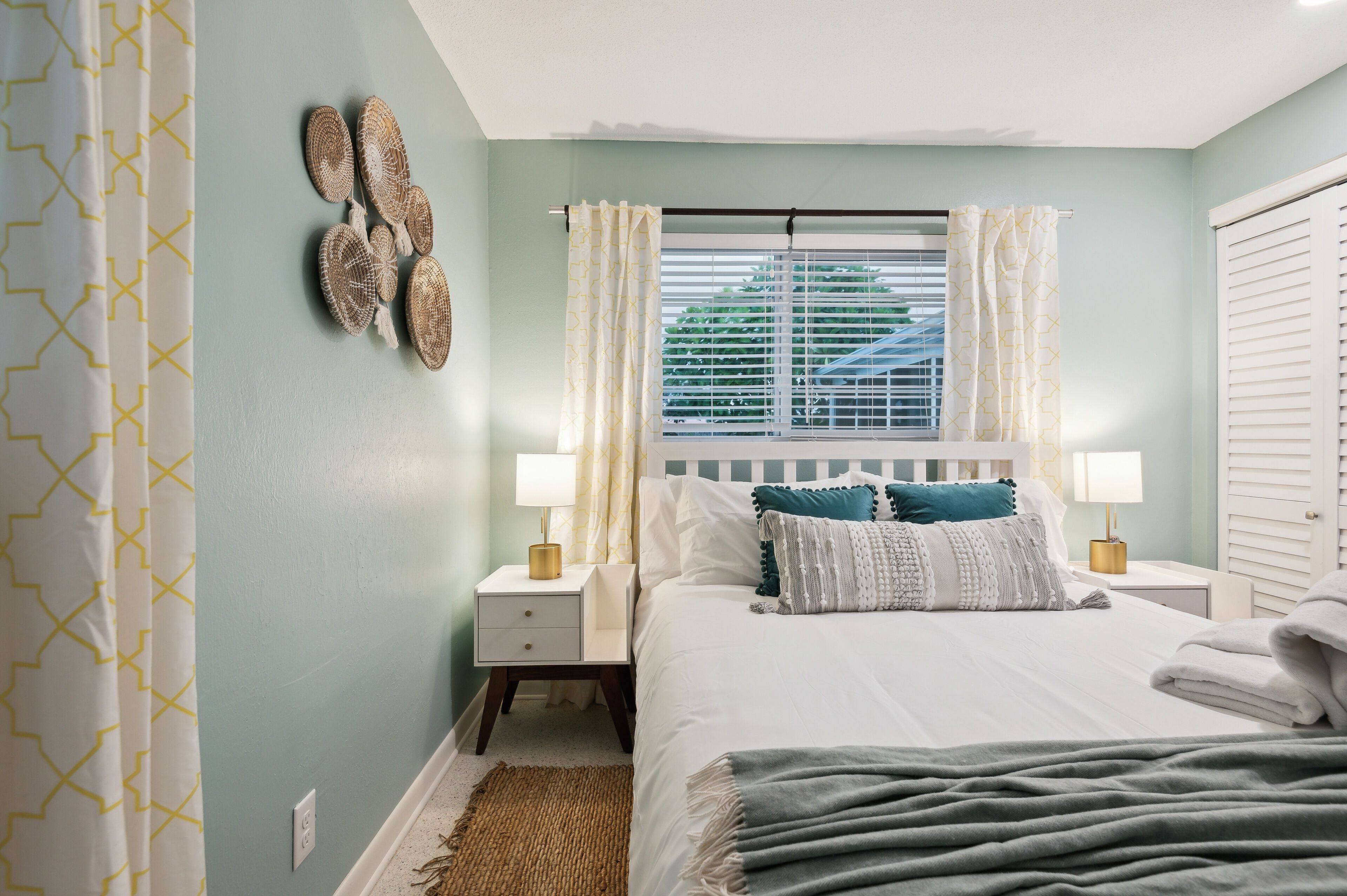 A bedroom with a bed, two bedside tables, a window with curtains, and a wall-mounted light fixture.