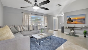 A modern living room with a grey sofa, a coffee table, a ceiling fan, and a large painting of a sunset over the ocean. - Boca Beauty - Stylish Home With Pool 3 Bedroom Home by RedAwning (Boca Raton)