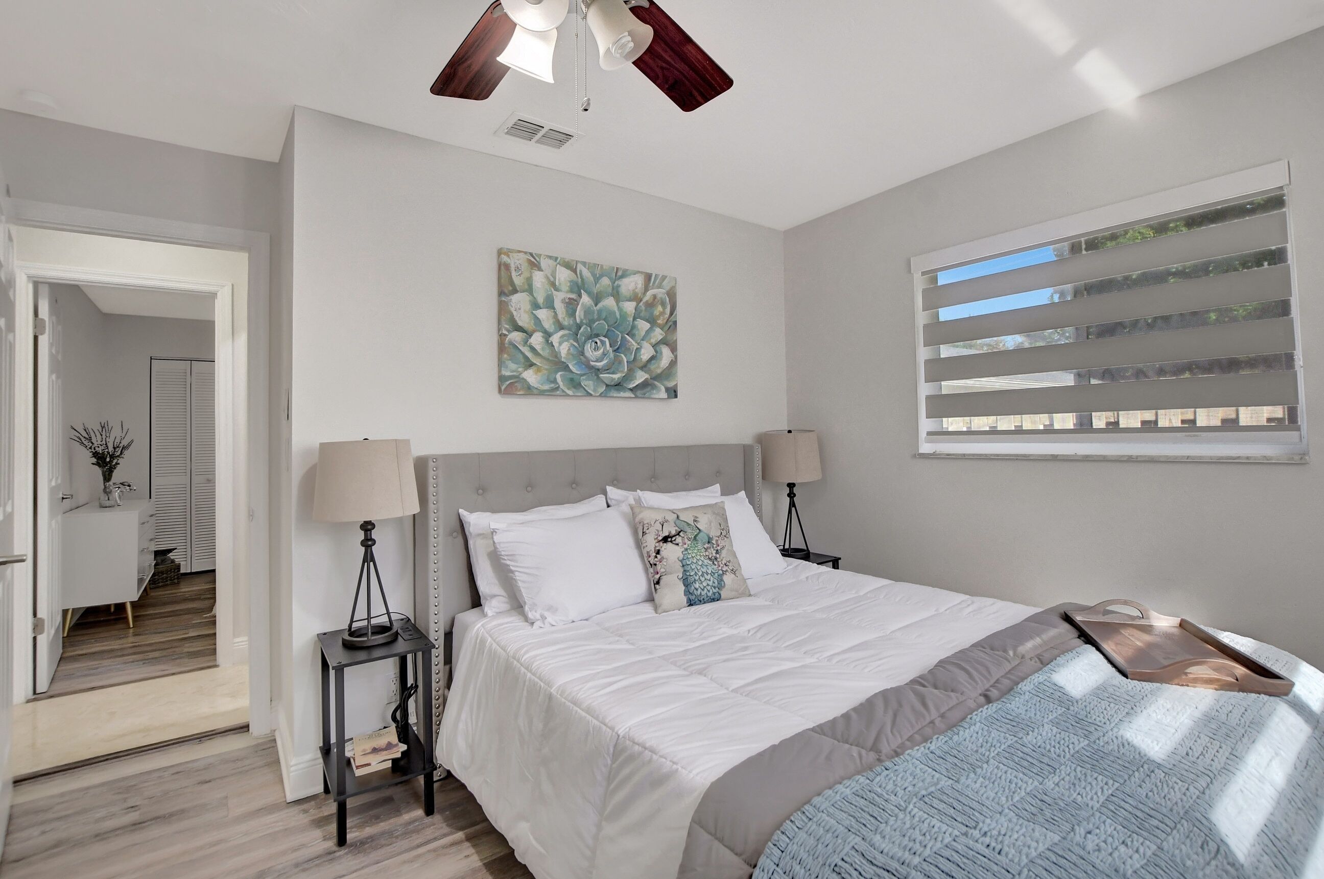 Un dormitorio con cama, mesitas de noche, ventilador de techo, ventana con persianas y puerta que da a otra habitación.