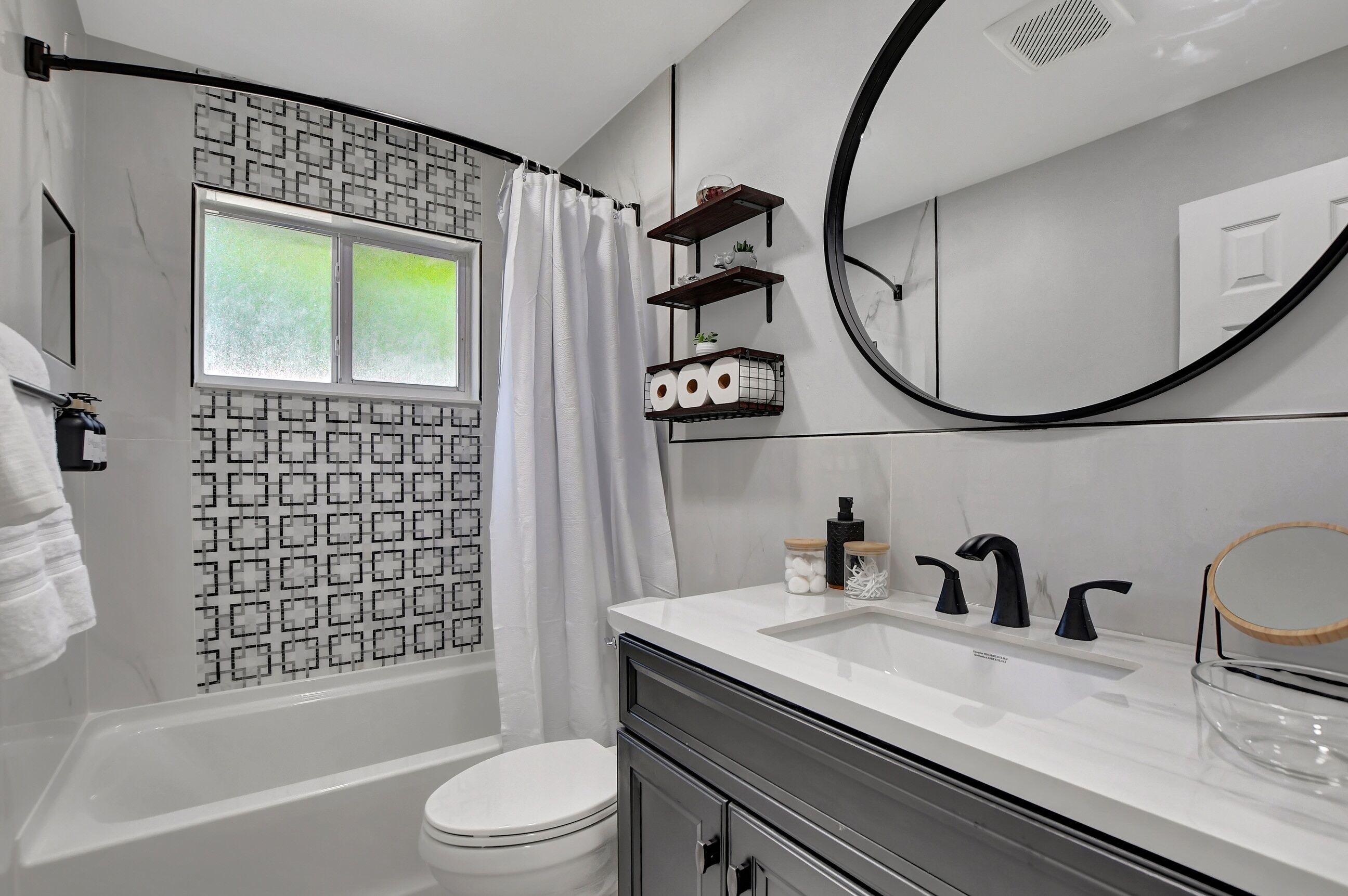 A modern bathroom with a bathtub, toilet, and sink. There is a window with a view of greenery, a shower curtain, and a mirror with a round frame.