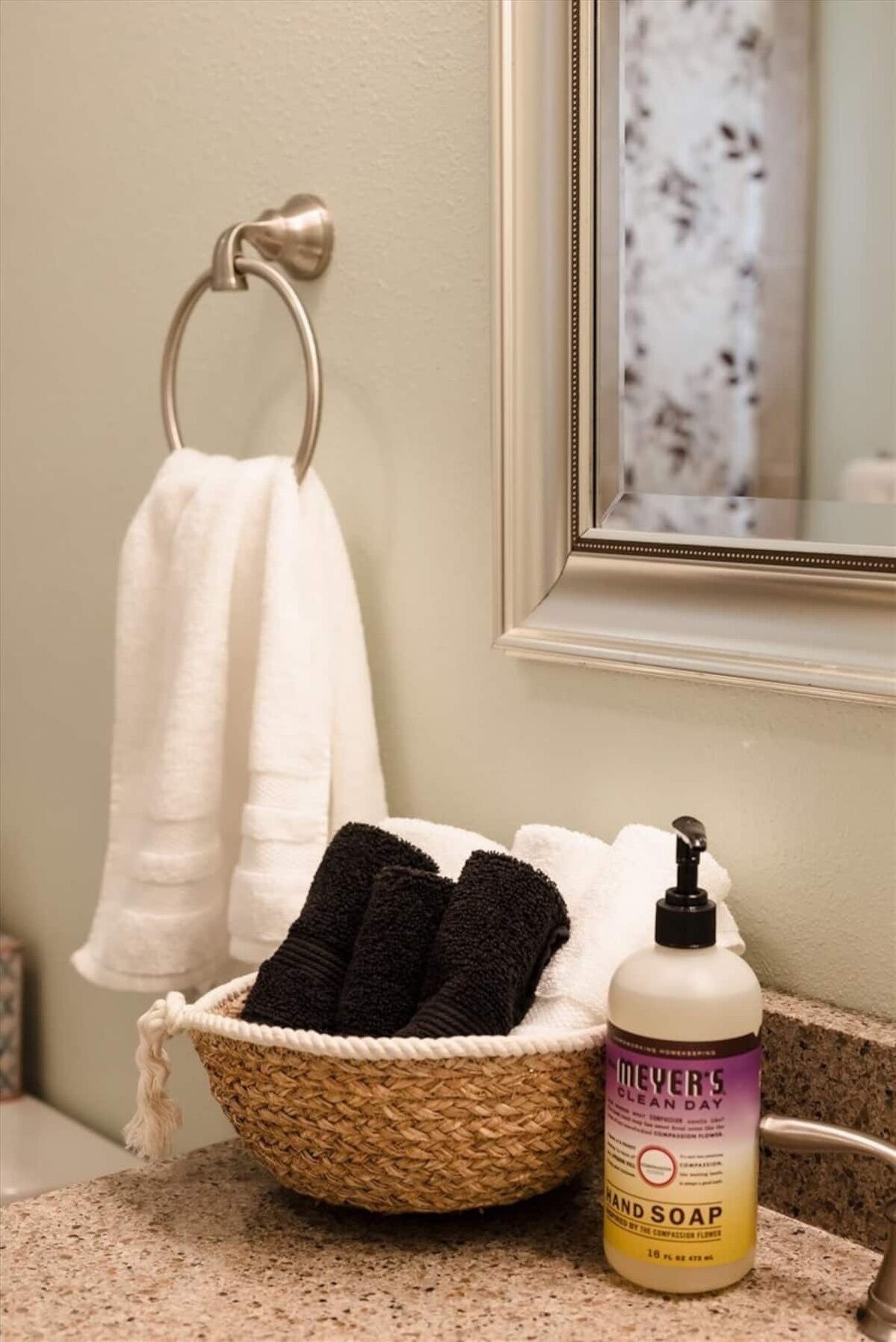 A bathroom with a white towel hanging on a metal towel rack, a wicker basket containing black and white towels, and a bottle of Meyer’s Clean Day hand soap on the countertop.