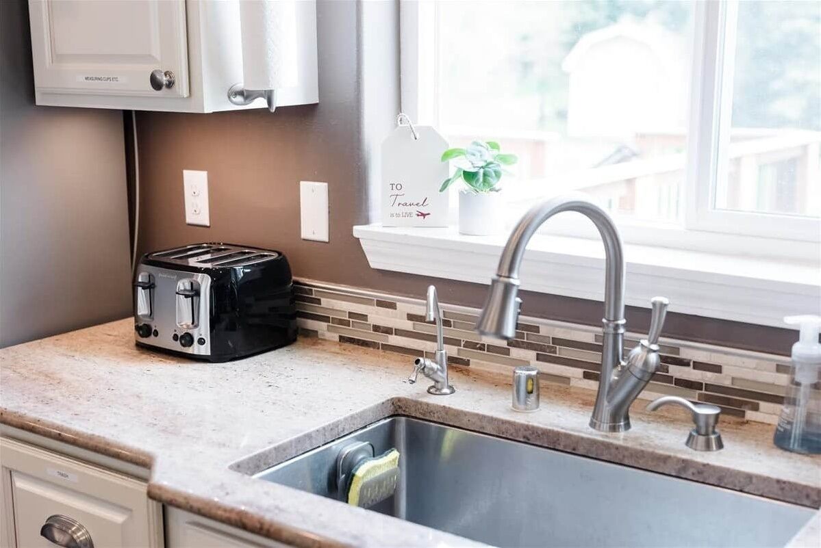 A kitchen with a double toaster, a sink with a modern faucet, a window with a plant, and a sign that reads "TO Travel".