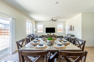 Comedor con una mesa grande dispuesta para seis personas, rodeada de sillas de madera, un ventilador de techo y un televisor montado en la pared.