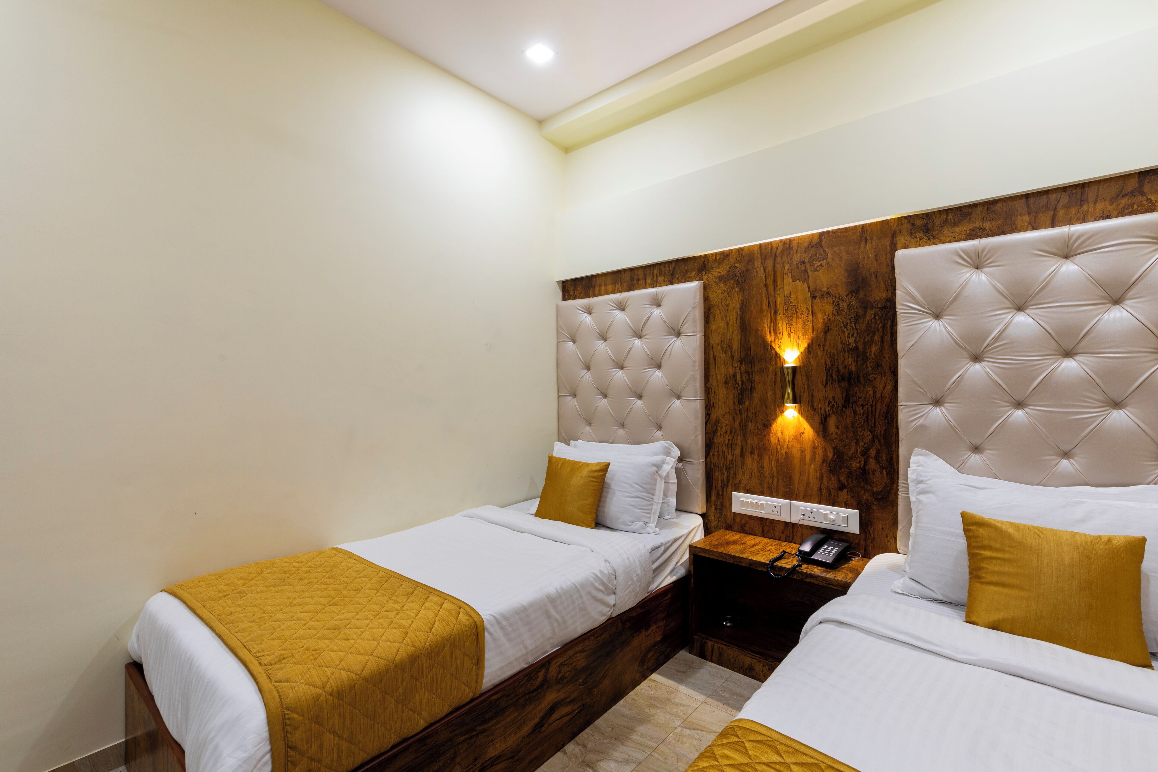 A hotel room with two beds, each with a wooden headboard, a bedside table with a phone, and a wall-mounted light fixture.