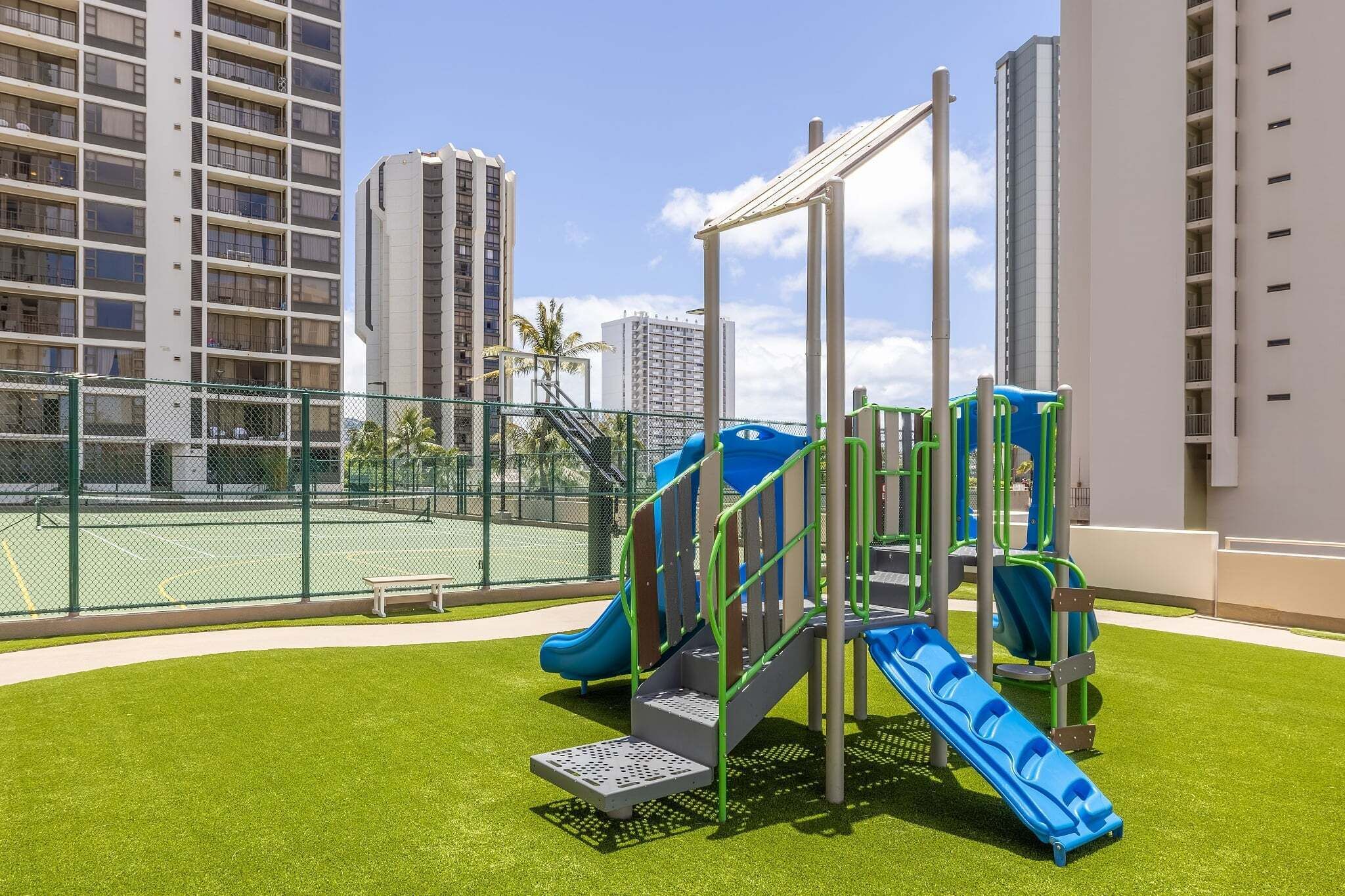 A playground with slides and climbing structures, surrounded by a tennis court and tall residential buildings.