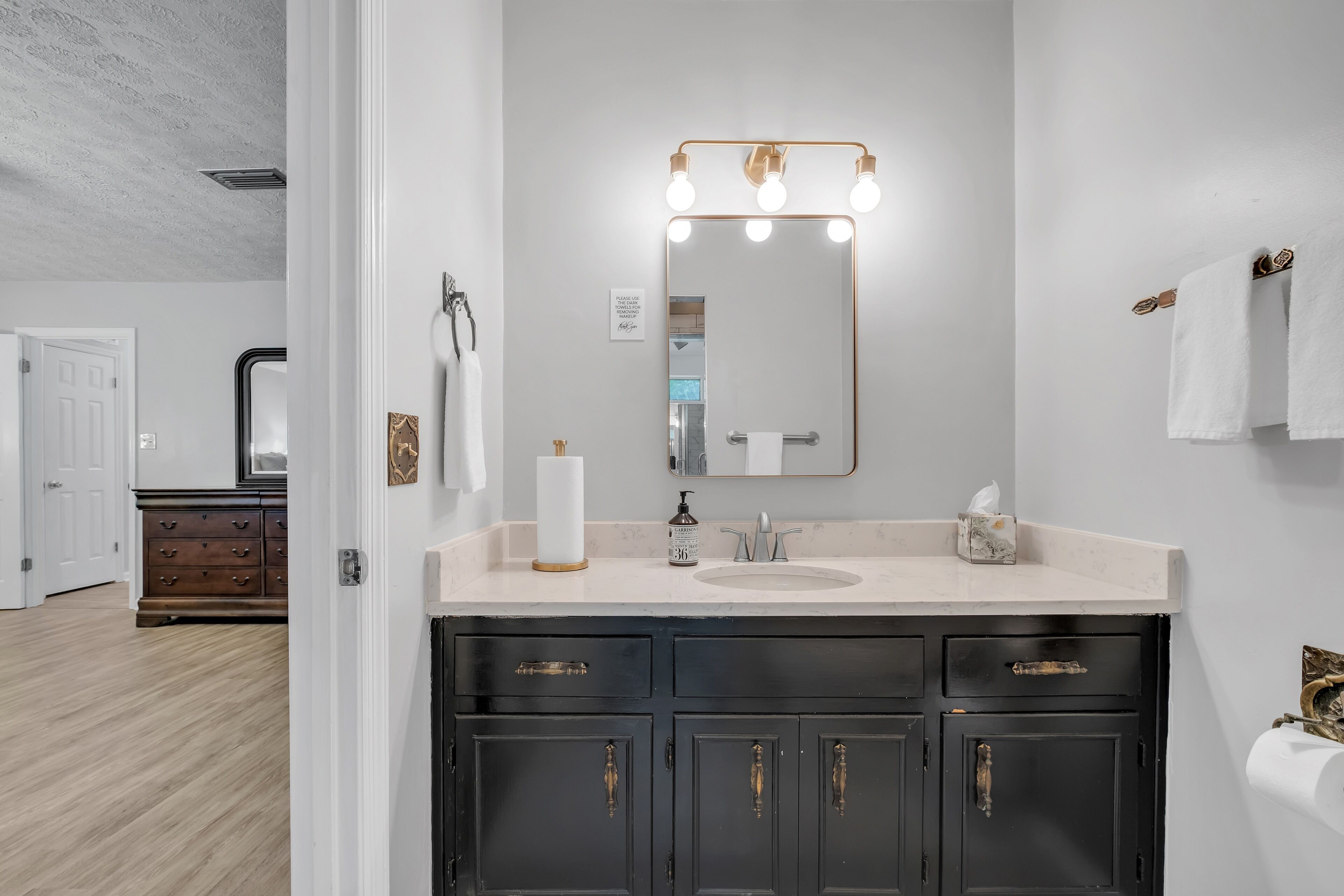 A bathroom with a vanity, a mirror with lights, a towel rack, and a toilet paper holder.