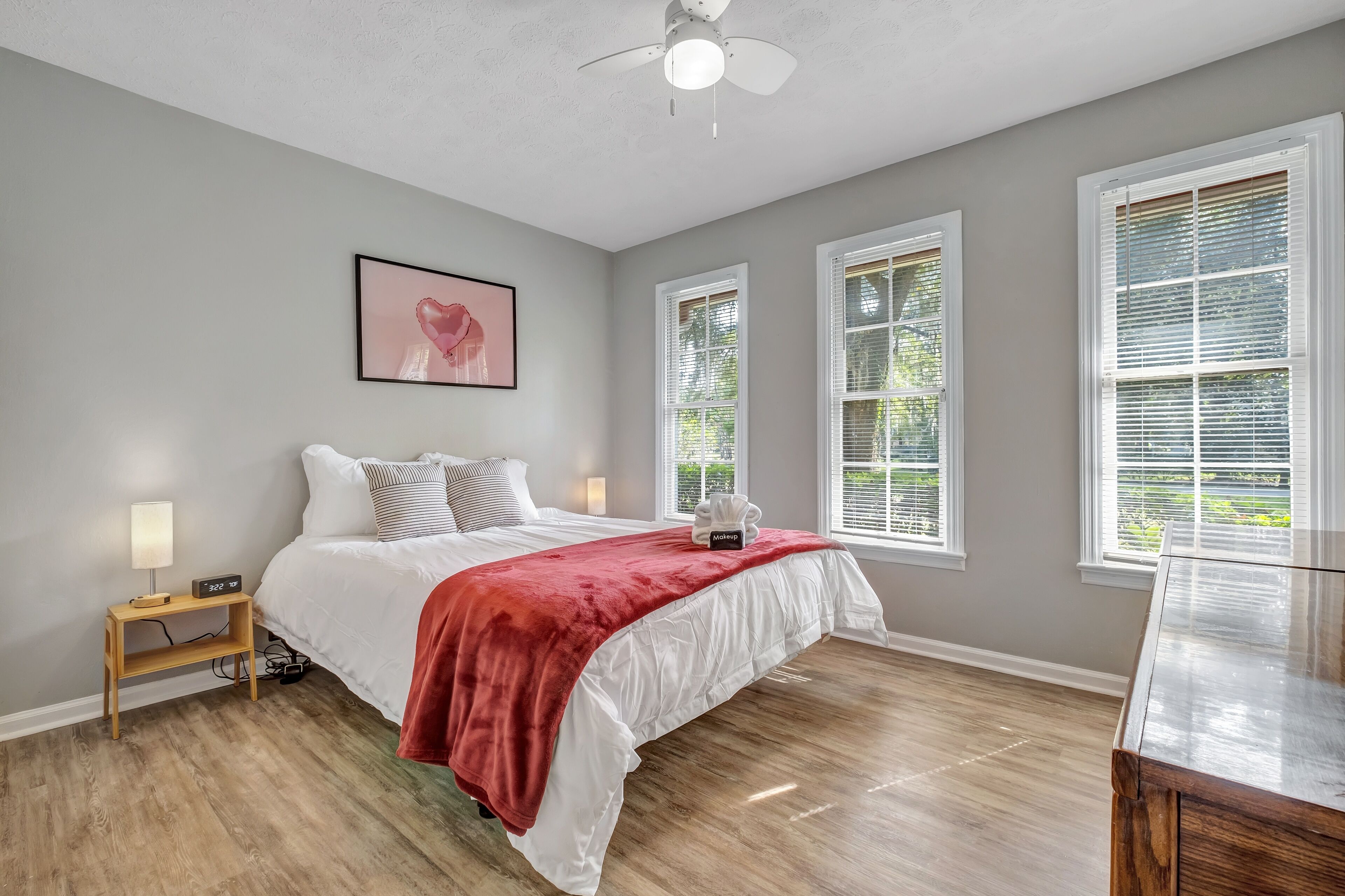 Une chambre à coucher avec un lit, une table de chevet, une lampe, un plaid rouge et un tableau encadré accroché au mur.