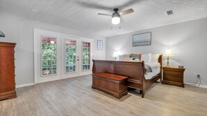 A bedroom with a wooden bed, a nightstand, a dresser, and a ceiling fan. There are two windows with red curtains and a view of greenery outside.