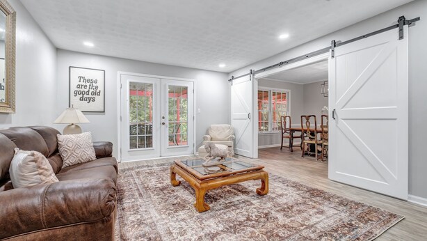 A spacious living room with a brown leather sofa, a glass coffee table, and a patterned rug. There is a sliding barn door leading to a dining area with a wooden table and chairs.