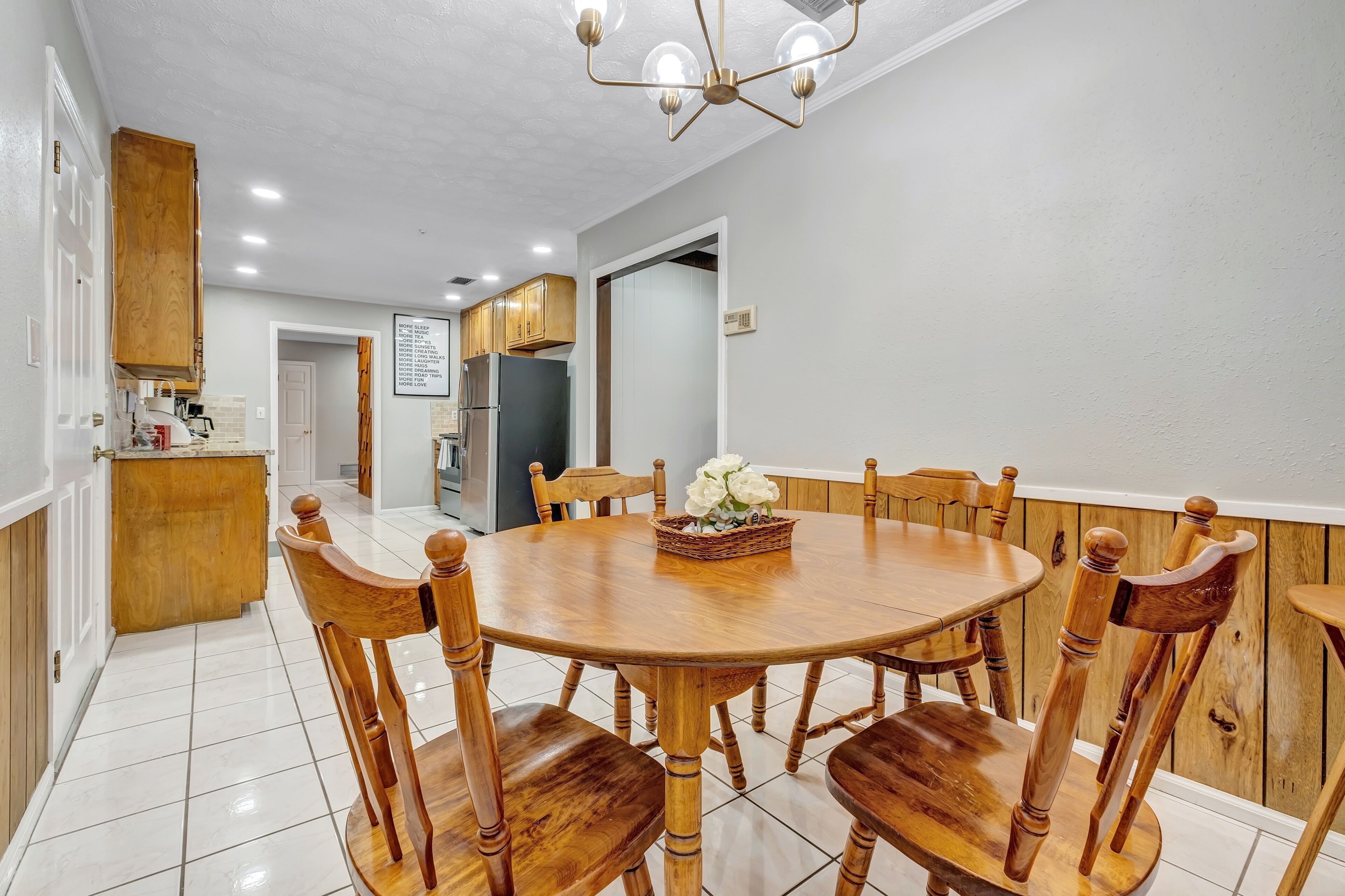 Un coin repas avec une table ronde en bois et des chaises, un lustre, et une cuisine en arrière-plan.