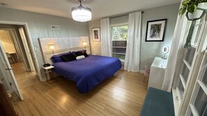 Ein Schlafzimmer mit einem großen Bett, Holzboden, einer Deckenleuchte, einem Fenster mit Vorhängen und einem Nachttisch mit Lampe.