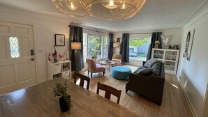 Ein geräumiges Wohnzimmer mit einem Holzesstisch, einem blauen Hocker, einem grauen Sofa und einem großen Fenster mit Blick ins Freie.