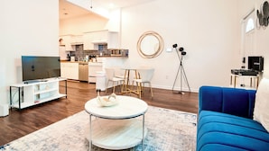 A modern living room with a blue sofa, a round coffee table, and a TV on a white stand. There is a kitchen in the background with white cabinets and a dining area with white chairs.