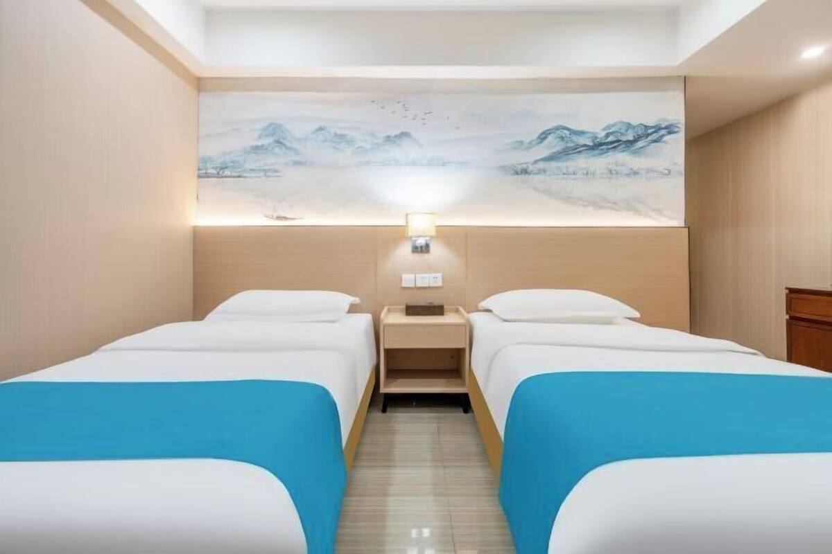 A hotel room with two beds, a nightstand, a wall-mounted lamp, and a large painting of a mountainous landscape.