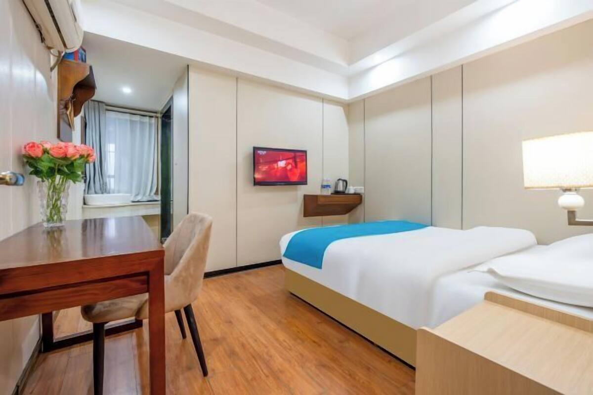 A hotel room with a wooden desk, a chair, a bed with a blue blanket, a television, and a bedside table with a lamp.