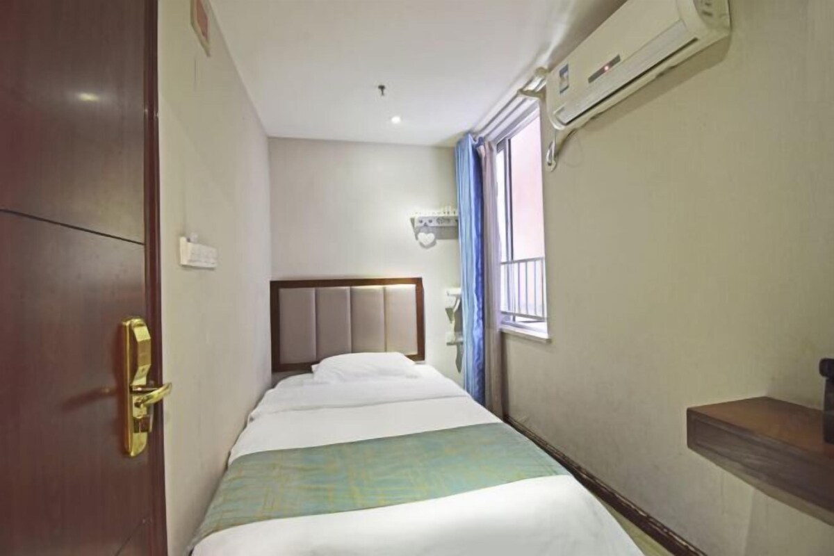 A narrow hotel room with a bed, a bedside table, a window with curtains, and an air conditioning unit.
