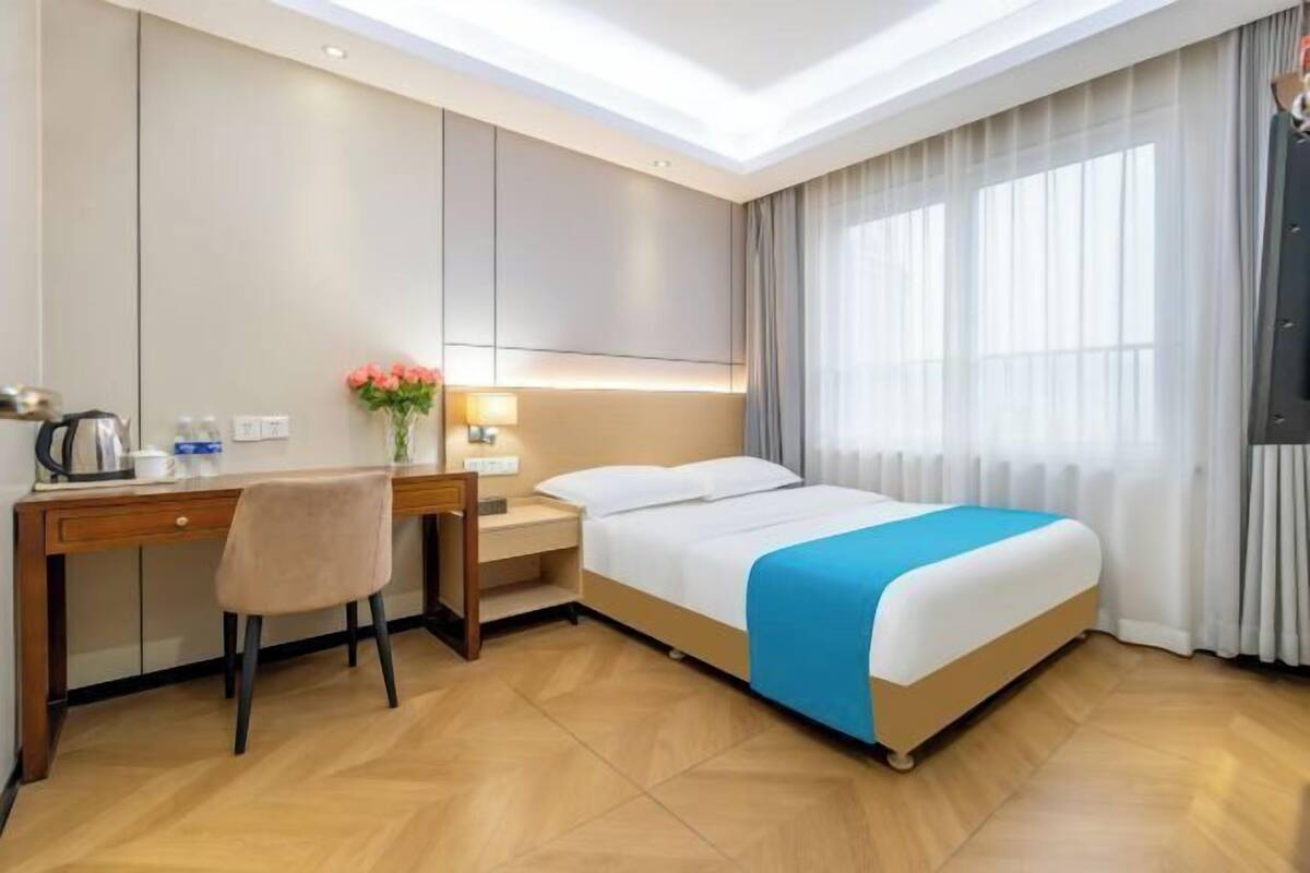 A hotel room with a bed, a desk, a chair, a nightstand, a lamp, a vase with flowers, and a large window with curtains.