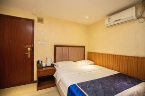 Habitación de hotel con puerta de madera, cama con cabecero, mesita de noche con hervidor y aire acondicionado en la pared.