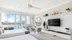 A modern hotel room with a large bed, a TV mounted on the wall, a dining area with a table and chairs, and a balcony with a city view. - EL SOL OCEAN VIEW / EXCLUSIVE  (San Juan)
