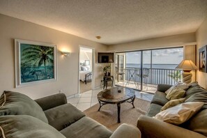 Ein Wohnzimmer mit einer Couch, einem Couchtisch und einem Fernseher. Es gibt einen Balkon mit Meerblick.