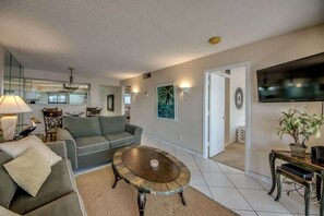 A living room with a sofa, coffee table, and a TV. There is a kitchen in the background.