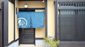 A traditional Japanese building entrance with a blue hanging banner, a black gate, and a potted plant. - Rinn Marikoji (Kyoto)