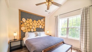 Une chambre à coucher avec un grand lit, une tête de lit en bois, un banc, un ventilateur de plafond, une fenêtre avec des rideaux et un luminaire fixé au mur.